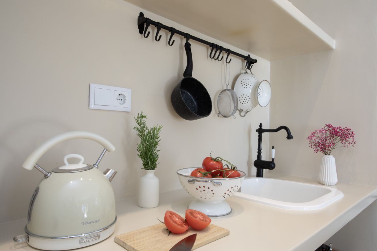 A kitchen with a kettle, colander, tomatoes, a cutting board, and a sink.