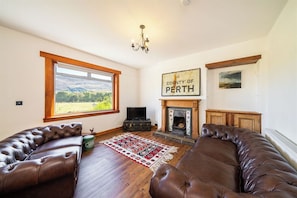 A living room with a fireplace, leather sofas, a television, and a large window with a view of the countryside. - Cosy Cottage close to Munros and Cycle path in Lochearnhead  (Lochearnhead)