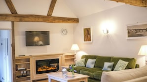 A living room with a fireplace, green sofa, white armchair, wooden beams, TV, wall clock, and artwork. - Broomhill Cottage (Petworth)