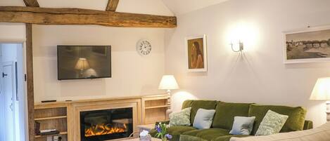 A living room with a fireplace, green sofa, white armchair, wooden beams, TV, wall clock, and artwork.