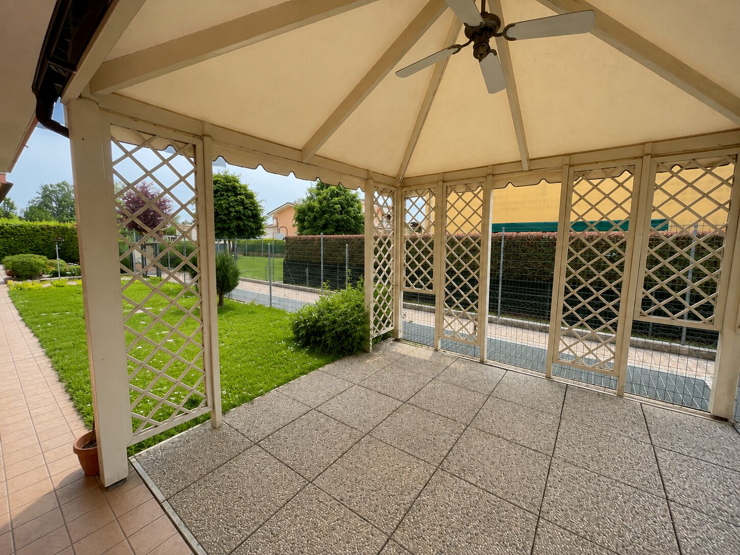 Un'area patio coperta con una ventola a soffitto, pavimento piastrellato e vista su un giardino curato e una recinzione.