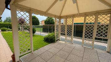 Un'area patio coperta con una ventola a soffitto, pavimento piastrellato e vista su un giardino curato e una recinzione.