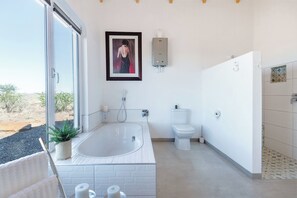 A modern bathroom with a large bathtub, toilet, and shower. There is a framed artwork on the wall and a potted plant on the counter. - A Luxurious Getaway in Namibia – Nature, Tranquility, and Endless Vastness (Maltahöhe)