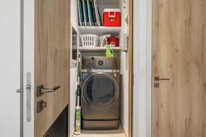 A compact laundry room with a front-loading washing machine, shelves with cleaning supplies, and a red storage box. - Walishali 215 (Oranjestad)