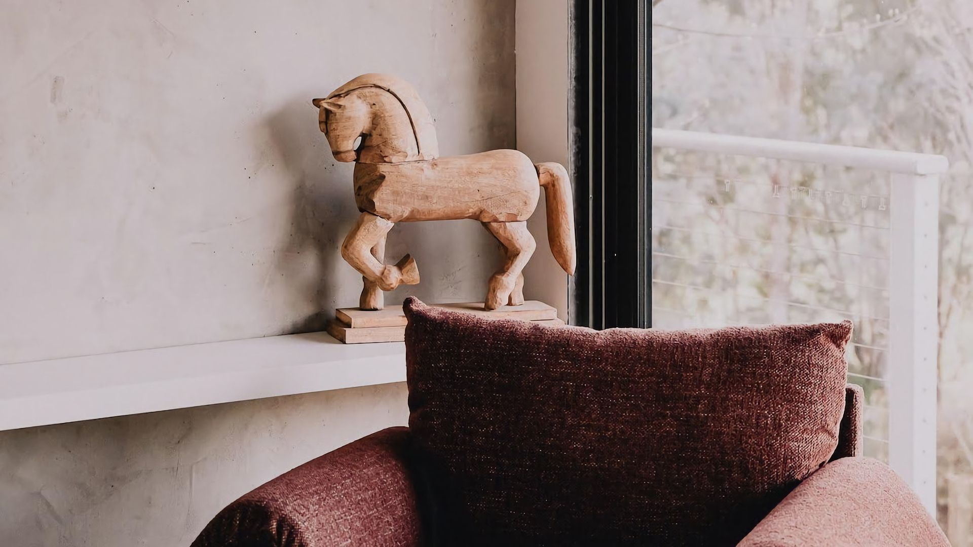 A wooden horse sculpture on a shelf, a window with a view of trees, and a dark-colored cushion on a chair.
