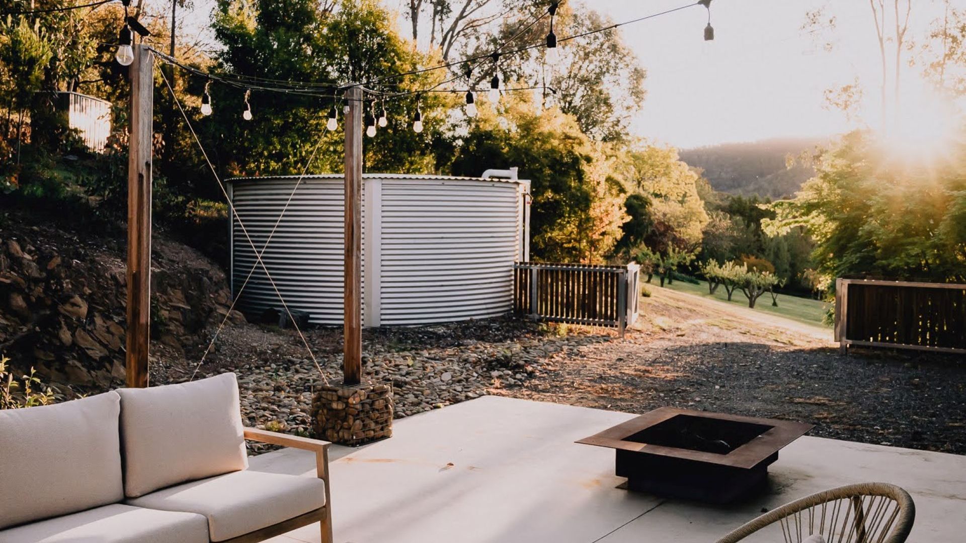 An outdoor seating area with a sofa and chairs, a fire pit, and a metal water tank in the background.