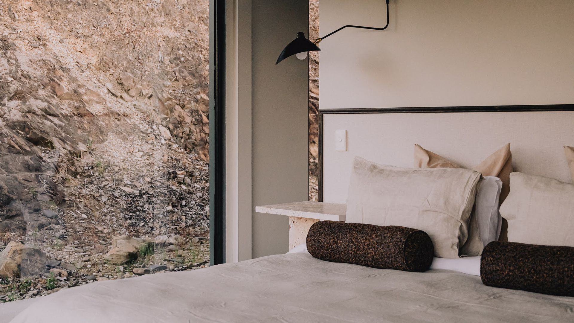 A bedroom with a stone wall, a bed with pillows, and a bedside lamp.