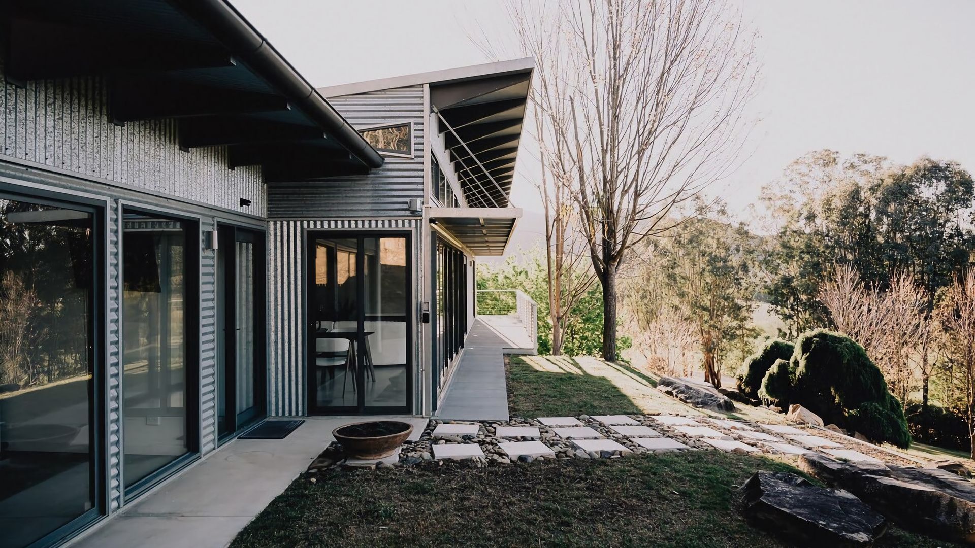A modern house with a glass facade, a covered outdoor seating area, and a stone pathway leading to the entrance.