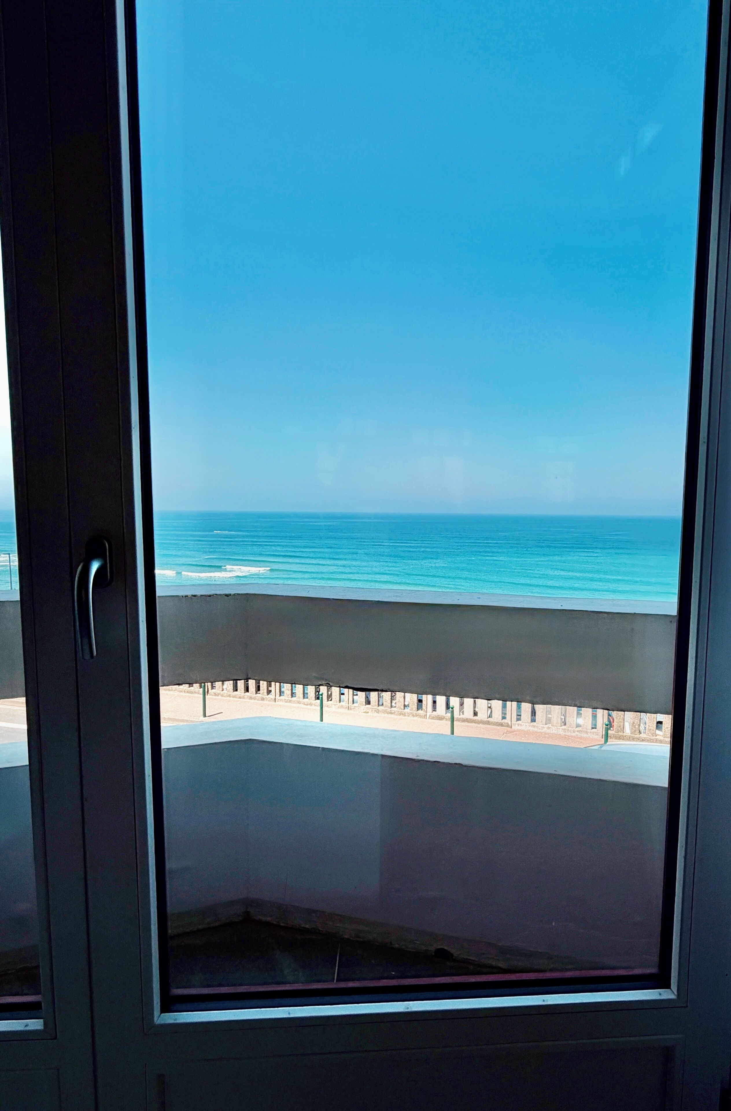 Vista desde una ventana a un balcón con vista a una playa y cielo despejado.