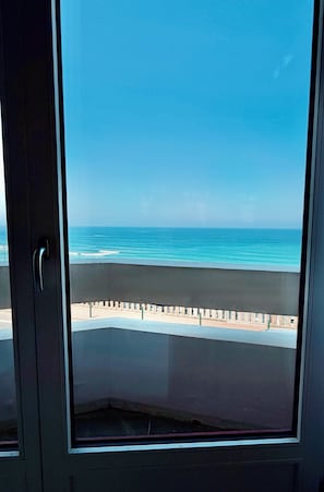 Blick durch ein Fenster auf einen Balkon mit Blick auf einen Strand und einen klaren Himmel.