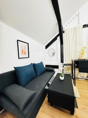 Ein modernes Wohnzimmer mit einem grauen Sofa, einem schwarzen Couchtisch, einem gerahmten Bild, einem runden Spiegel und einem kleinen Tisch mit Blumen.