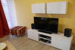 A living room with a flat-screen TV on a white cabinet, a red curtain, and a wooden floor. - Holiday apartment with 2 bedrooms (Walsrode)