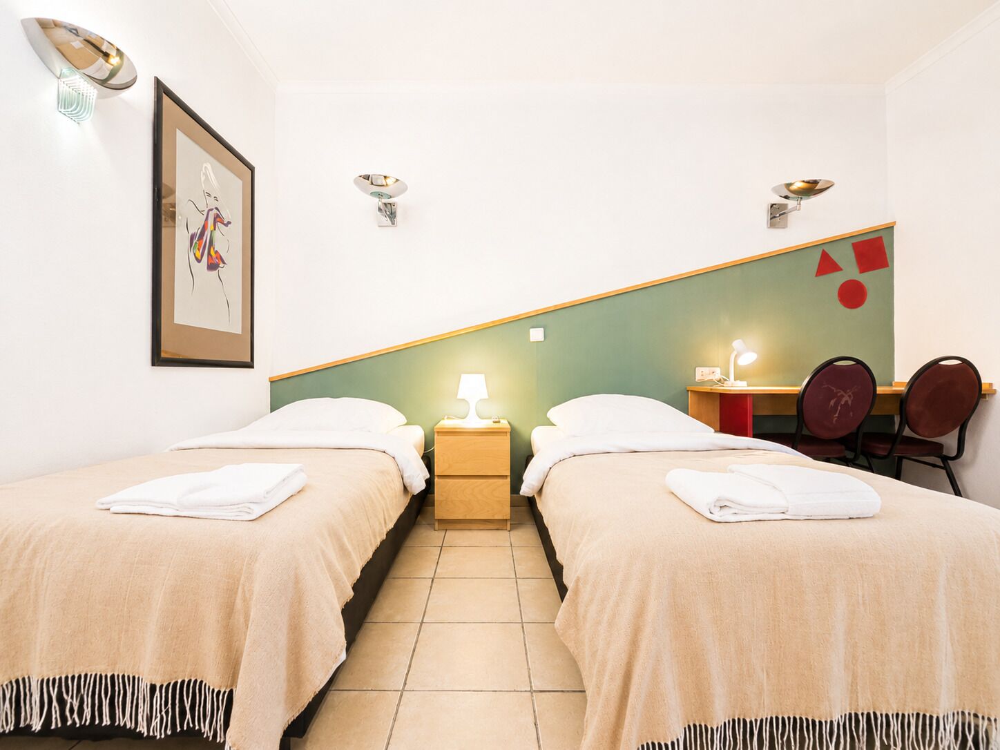 Habitación de hotel con dos camas, un escritorio, una silla, una lámpara y un cuadro en la pared.