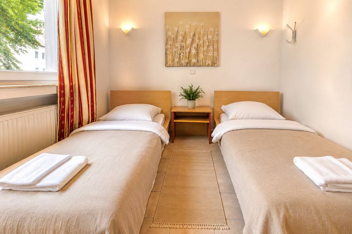A hotel room with two beds, a window with curtains, a painting on the wall, and a small table with a plant.
