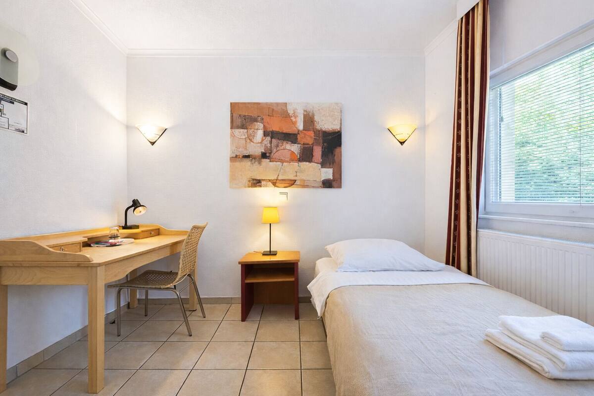 A hotel room with a bed, a desk, a chair, a lamp, and a painting on the wall.