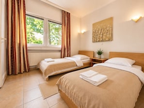 Ein Hotelzimmer mit zwei Betten, einem Fenster mit Vorhängen, einer Wandleuchte und einer Pflanze auf einem Nachttisch.