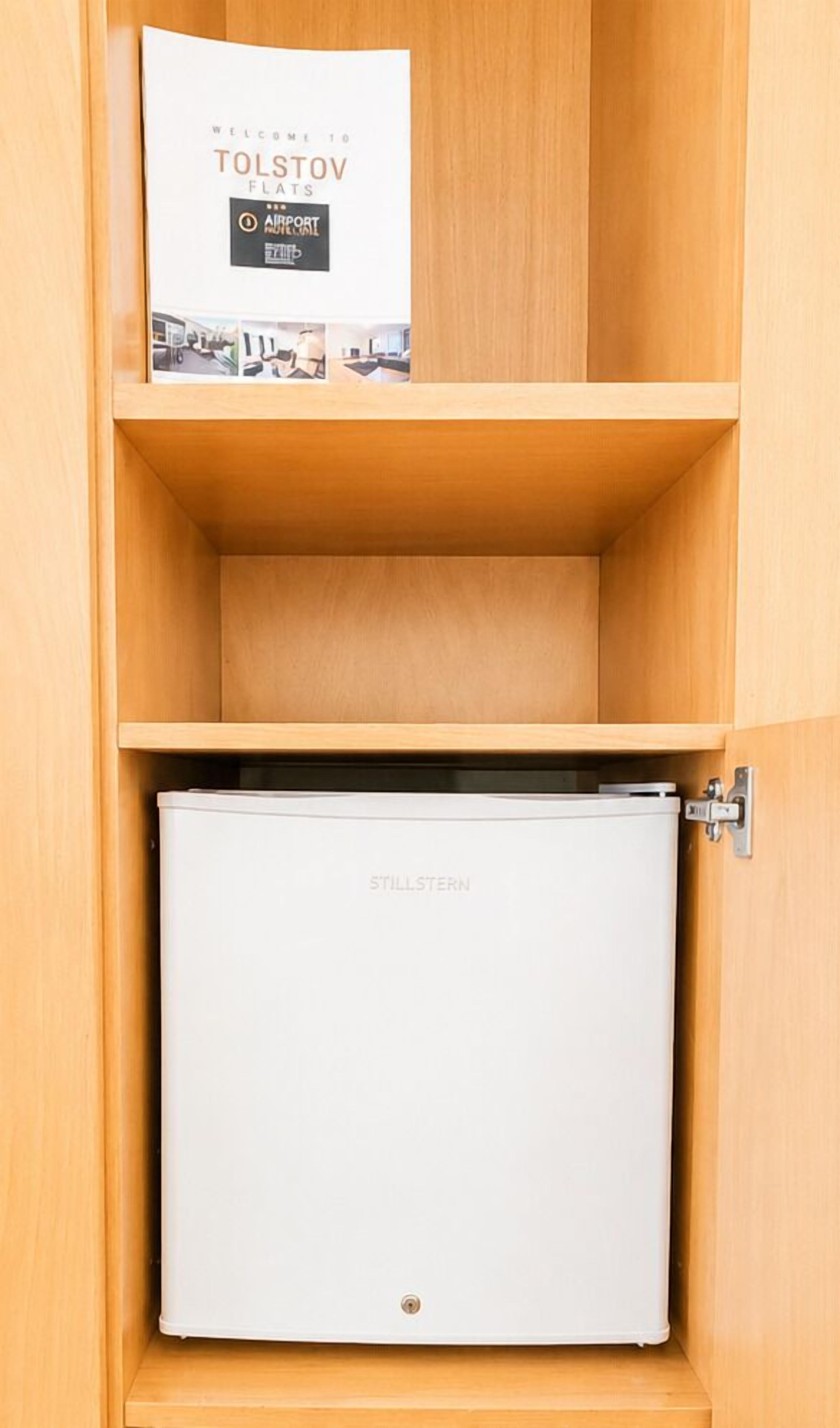 A wooden cabinet with a brochure titled 'Welcome to Tolstov Flats' and a white Stillstern refrigerator.