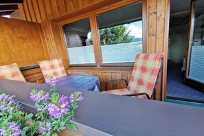 Interior de una cabaña de madera con sillas de cuadros, una mesa y un ventanal con vistas a árboles.