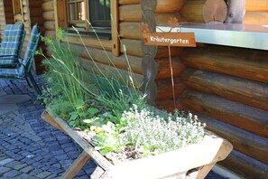 Ein Holzscherenkarren mit einem Schild, auf dem 'Kräutergarten' steht, neben einer Blockhütte mit Fenster und einem Stuhl davor.