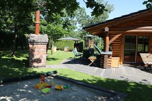 Eine Blockhütte mit Kamin aus Ziegelsteinen, Sitzgelegenheiten im Freien und einem Spielbereich mit Rutsche und Schaukeln.