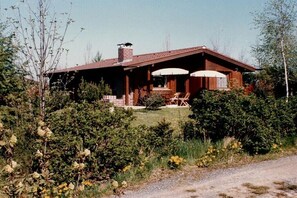 Ein einstöckiges Haus mit einem Ziegelkamin, einer Veranda mit Tisch und Stühlen sowie einem kleinen Garten.