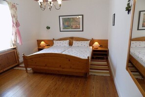 Ein hölzernes Bett mit zwei Nachttischen, einem Heizkörper, einem Fenster mit Vorhängen und einem gerahmten Bild an der Wand.