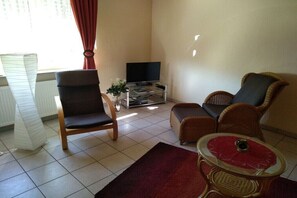 Ein Wohnzimmer mit Fernseher, zwei Korbstühlen, einem kleinen Tisch und einem roten Teppich.