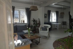 Un salon comprenant un canapé, un fauteuil en osier, une petite table avec un vase et une fenêtre avec des rideaux.