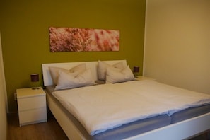 A single bed with a white mattress and pillows, a nightstand with a lamp, and a painting on the wall. - Holiday apartment with 2 bedrooms (Walsrode)