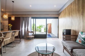 Ein modernes Wohnzimmer mit Glastisch, einer Couch mit tropischem Kissen, Korbstühlen, einer Küchenzeile mit Esstisch und einer Glastür, die auf einen Balkon führt.