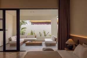 A modern hotel room with a bed, two sofas, a coffee table, and a view of a balcony with plants and a thatched roof.