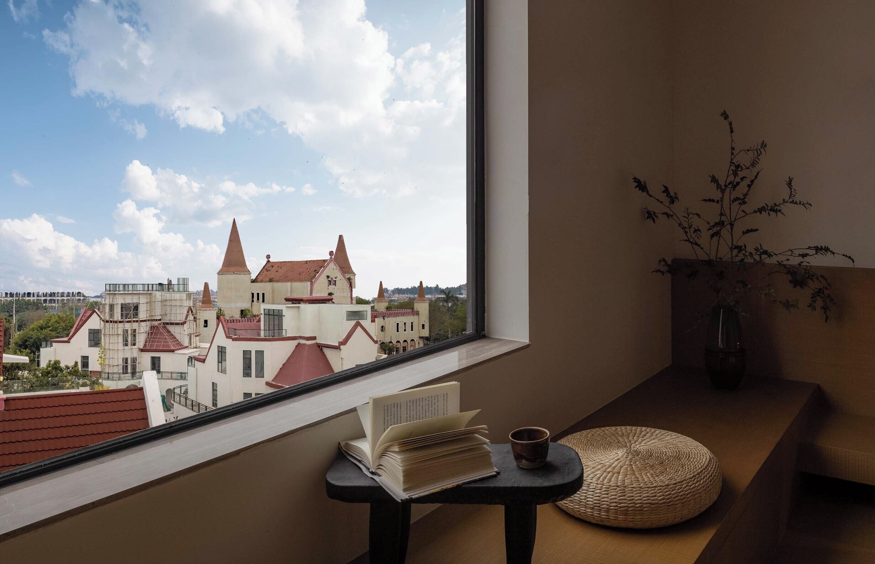 A room with a view of a castle, a table with a book and a cup, and a vase with branches.