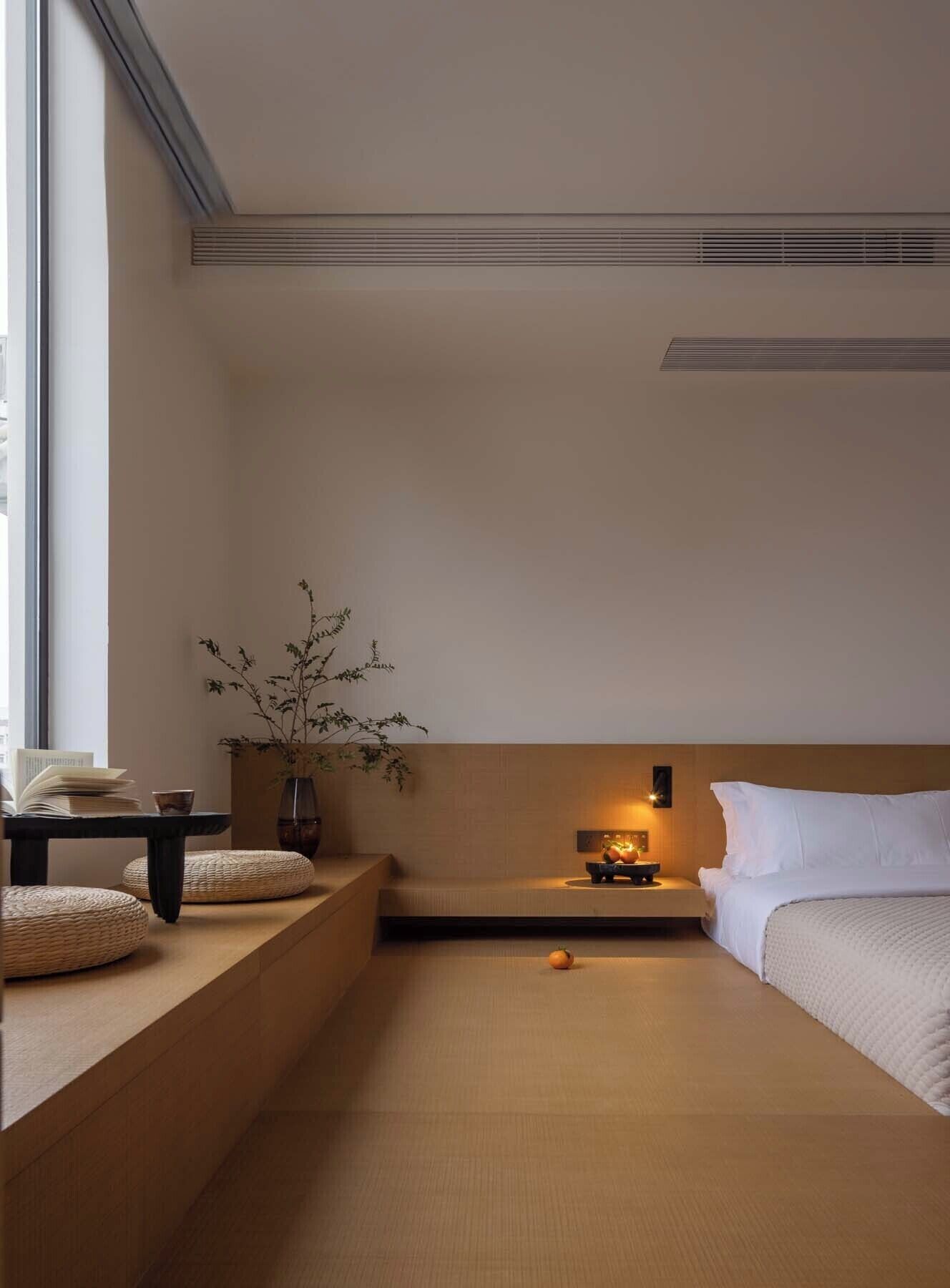 A minimalist room with a bed, a small table with books, a vase with branches, and a nightstand with a lamp.