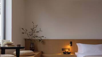 A minimalist room with a bed, a small table with books, a vase with branches, and a nightstand with a lamp.
