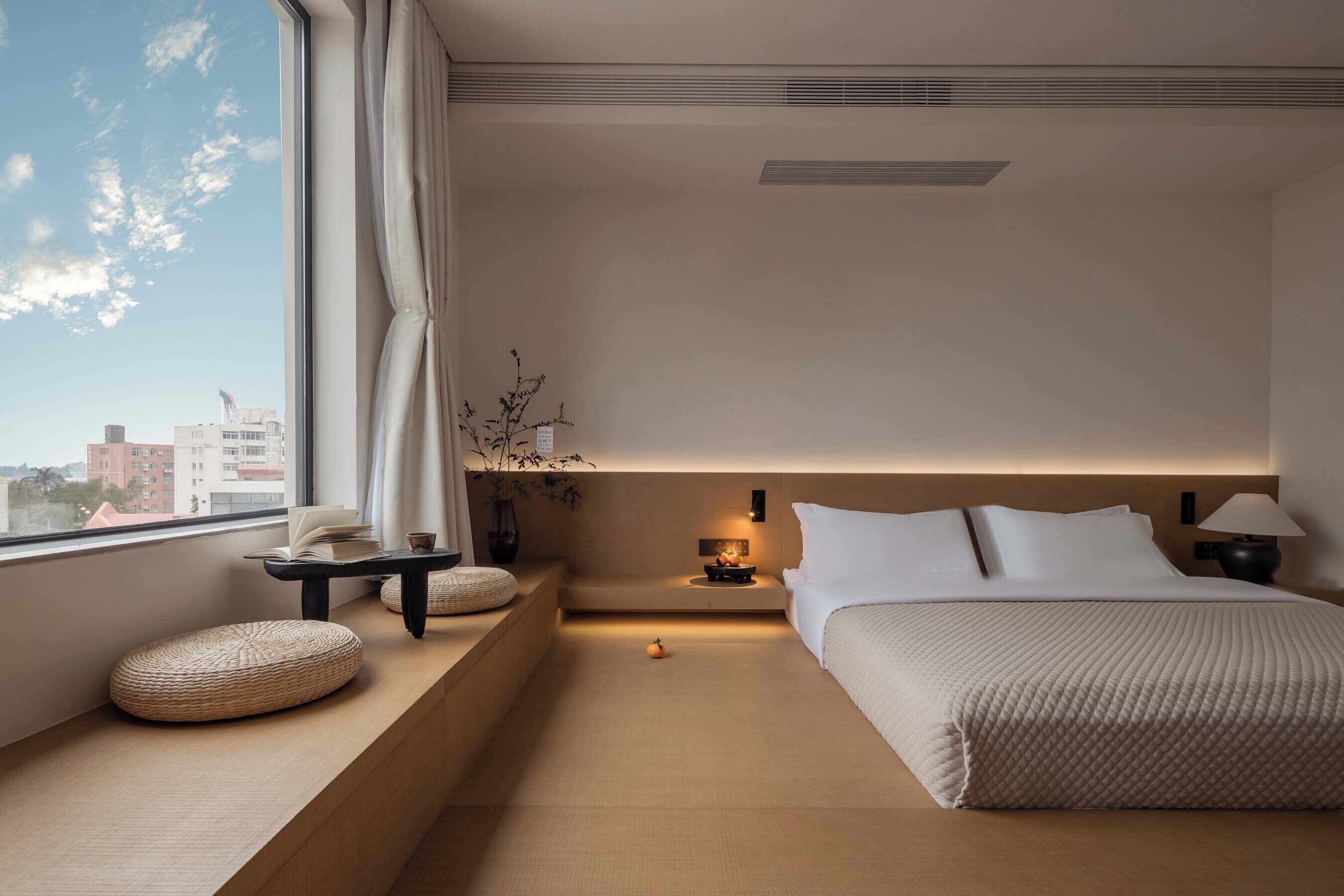 Une chambre d’hôtel moderne avec une grande fenêtre, une table de chevet avec une lampe, un lit avec une literie blanche et une petite plante sur le rebord de la fenêtre.