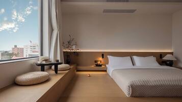 Une chambre d’hôtel moderne avec une grande fenêtre, une table de chevet avec une lampe, un lit avec une literie blanche et une petite plante sur le rebord de la fenêtre.