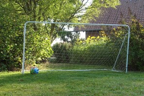 Ein Fußballtor auf einer Rasenfläche mit Netz, umgeben von Grünpflanzen und einem Gebäude im Hintergrund.
