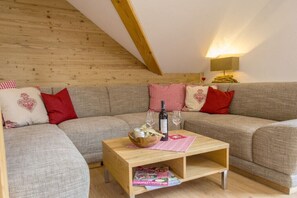 Ein gemütliches Wohnzimmer mit Holzbalkendecke, einer Sofagarnitur, einem Couchtisch mit Flasche und Gläsern sowie einer Lampe.