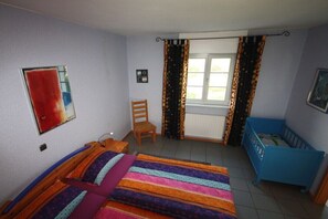 Ein Schlafzimmer mit einem Bett, einem Sessel, einem blauen Babybett, einem Fenster mit Vorhängen und einer Heizung.