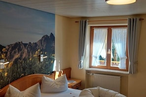 Ein Hotelzimmer mit einem Bett, einem Nachttisch, einem Fenster mit Vorhängen und einem Wandbild von einer Berglandschaft.