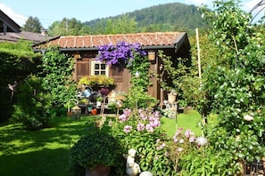 A rustic wooden house with a tiled roof, surrounded by lush greenery and flowering plants. - Holiday apartment with 1 bedroom (Bad Wiessee)