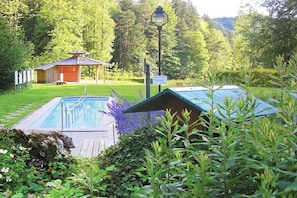 Ein Schwimmbecken mit Holzterrasse, einer Laube und einer Laterne, eingebettet in eine üppige grüne Umgebung.