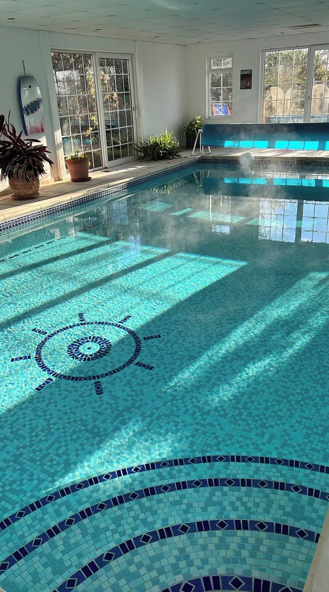 Une piscine intérieure à l’eau d’un bleu cristallin, dont le sol est décoré d’un motif de cadran solaire, et des plantes en pot qui ornent l’espace autour de la piscine.