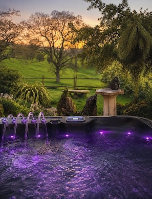 A hot tub with a stone pedestal and a butterfly on it, surrounded by greenery and a sunset in the background.