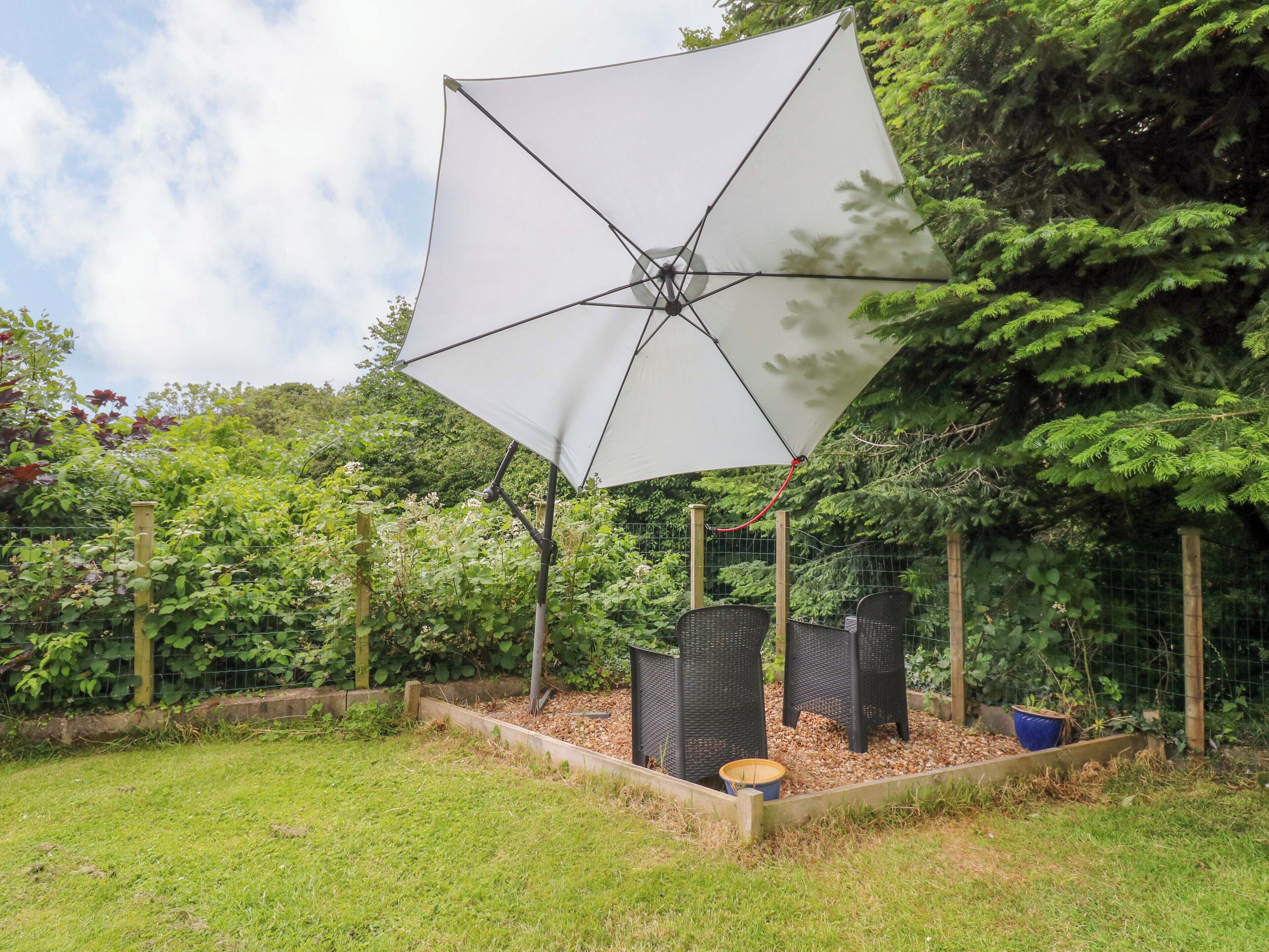 An outdoor area with a large white umbrella, wooden chairs, and a garden bed with plants.