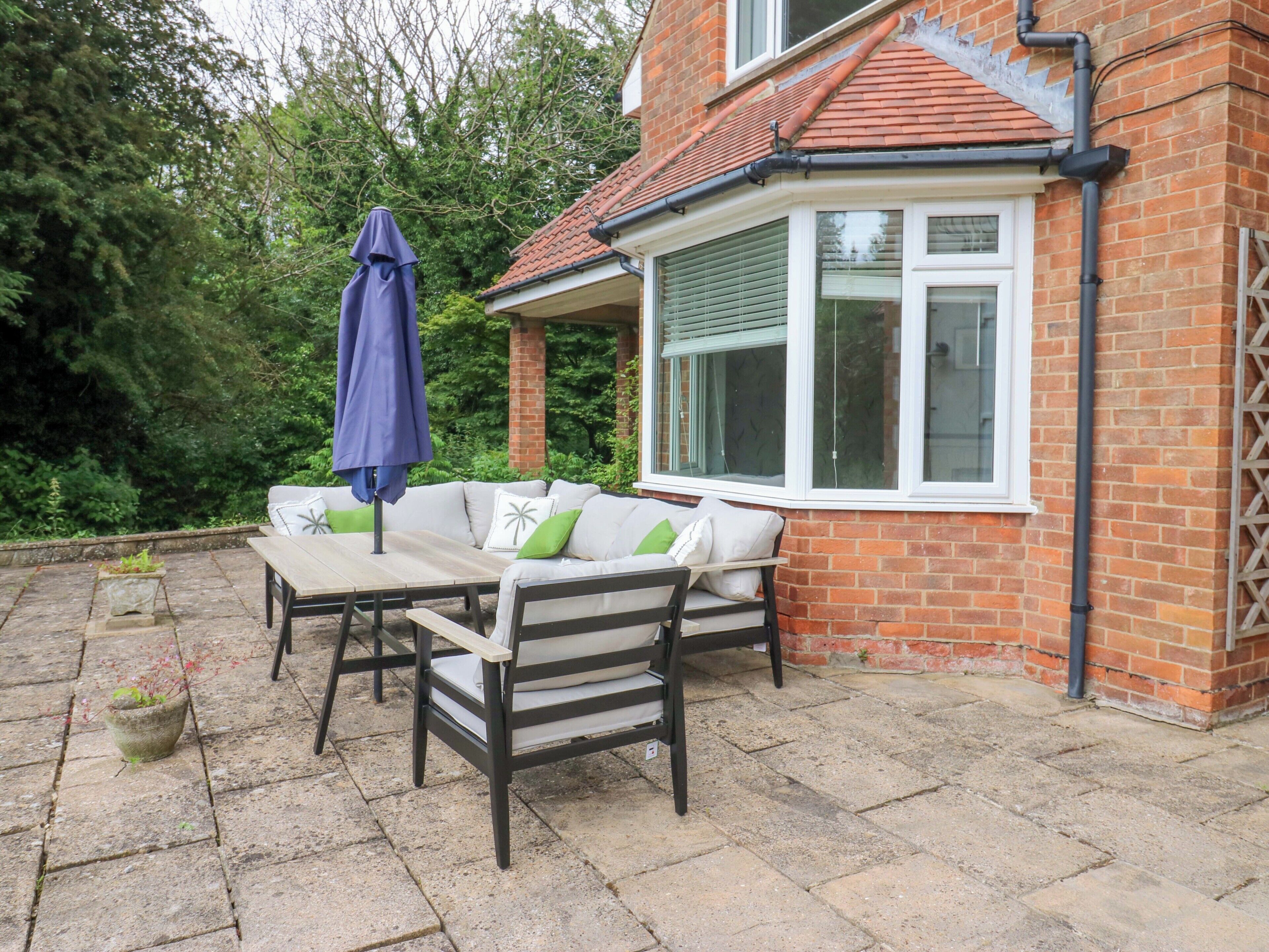 A patio with a table and chairs, a blue umbrella, and a brick house with a bay window.