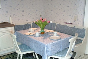 Ein Esstisch mit einer blumigen Tischdecke, weißen Stühlen und einer Vase mit rosafarbenen Blumen.