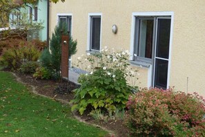 Ein Gebäude mit drei Fenstern, ein Garten mit unterschiedlichen Pflanzen und ein Weg zum Eingang.