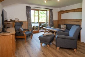 Ein gemütliches Wohnzimmer mit Holzboden, einem Fernseher, einem Sofa, einem Sessel, einem Tisch und einem Fenster mit Vorhängen.