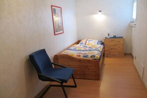 Ein kleines Schlafzimmer mit einem Einzelbett, einem Sessel, einer Kommode und einem gerahmten Bild an der Wand.